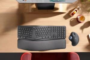 Top view of a modern workspace with a black ergonomic keyboard with wrist support, a vertical mouse, and a monitor on a wooden desk, alongside a cup of tea with a lemon slice.