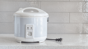 A simple white rice cooker on a speckled countertop with a modern tiled backsplash, plugged out and ready for use.