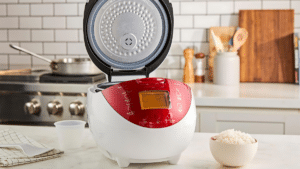 A white rice cooker with a red digital display, lid open, on a kitchen counter beside a bowl of cooked rice.