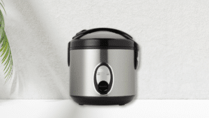 Stainless steel rice cooker with a black lid and simple cook/warm switch, placed against a textured white wall.