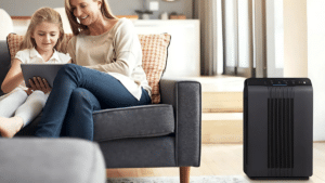 A mother and daughter sit on a gray couch in a cozy living room, with the mother smiling as the daughter uses a tablet. A black air purifier stands nearby, blending into the modern, light-toned decor.