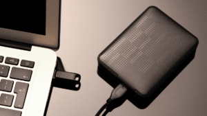 External hard drive connected to a laptop, with a USB flash drive plugged into the laptop, placed on a reflective surface.