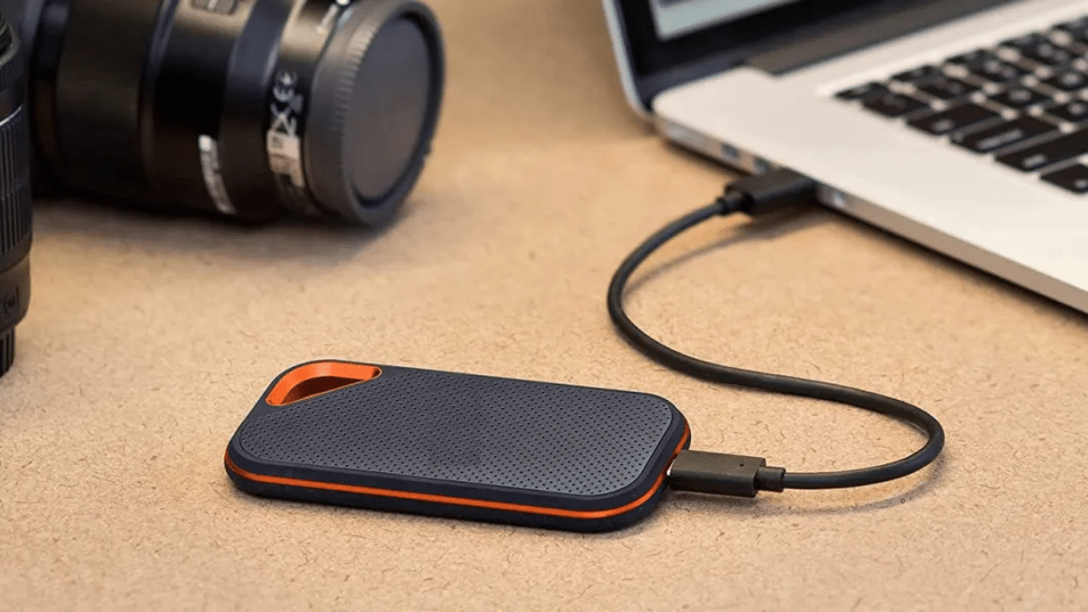 A compact external hard drive connected to a laptop, with a camera lens in the background, suggesting a setup for photographers or content creators needing portable storage.