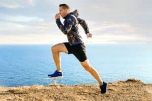 Man running along a coastal path, wearing a camouflage jacket and blue sneakers with the ocean in the background.